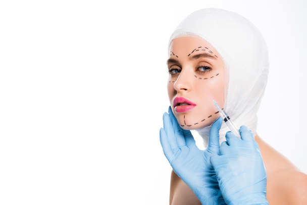 vista ritagliata del chirurgo plastico in guanti di lattice tenuta siringa vicino al viso della giovane donna con segni isolati su bianco
  - Foto, immagini