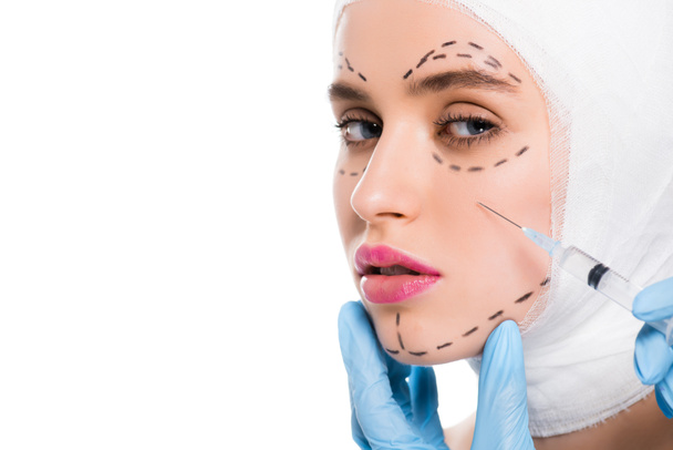 vista recortada del cirujano plástico en guantes de látex que sostiene la jeringa cerca de la cara de la mujer con marcas aisladas en blanco
  - Foto, Imagen