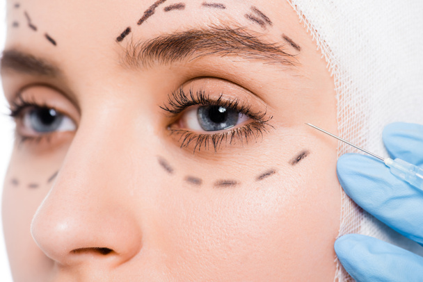 wykadrowany widok chirurga plastycznego w rękawiczce lateksowej w pobliżu twarzy kobiety ze znakami na białym  - Zdjęcie, obraz