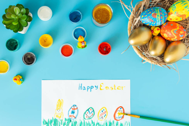 Easter concept, basket with hand made Easter eggs next to multicolor paints brush and white paper with sign happy Easter and paint multicolor Easter eggs on a blue bacground - Фото, изображение