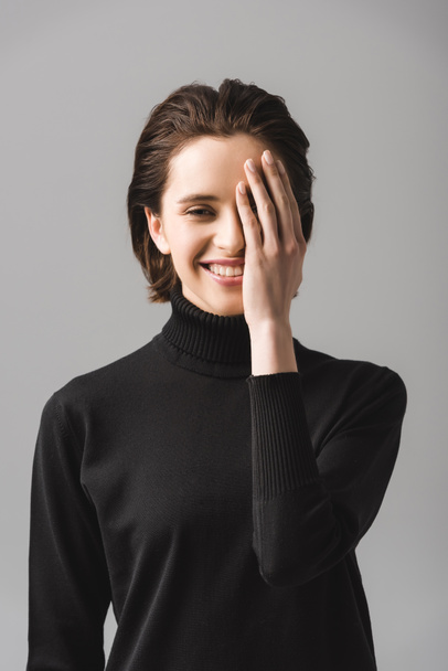 heureuse jeune femme en pull noir couvrant oeil isolé sur gris
  - Photo, image