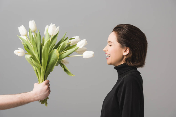 abgeschnittene Ansicht eines Mannes mit weißen Tulpen in der Nähe einer fröhlichen Frau isoliert auf grau  - Foto, Bild