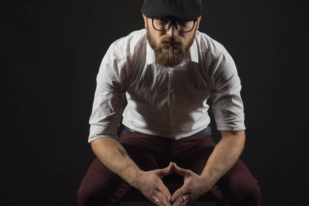 young pretty big bearded man in glasses with a beard and a mustache in a black cap and a white shirt sits on a chair looking directly seriously and purposefully, a dark studio background - Photo, Image