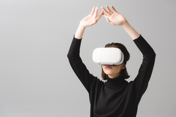 brunette woman wearing virtual reality headset while standing with hands above head isolated on grey  - Foto, immagini