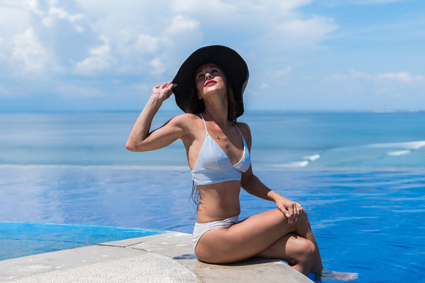 Привлекательная женщина в бикини и в серой шляпе расслабляется в бесконечном бассейне
 - Фото, изображение