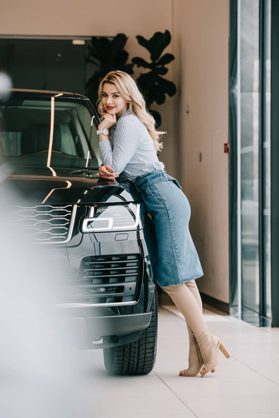selective focus of happy blonde girl standing near black automobile in car showroom   - Fotografie, Obrázek