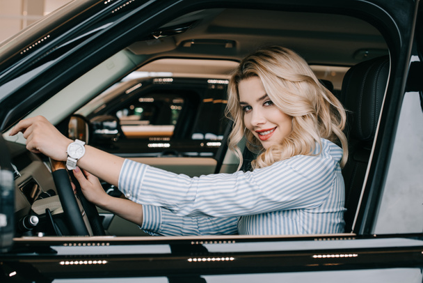 cheerful blonde woman holding steering wheel while sitting in car  - Fotó, kép