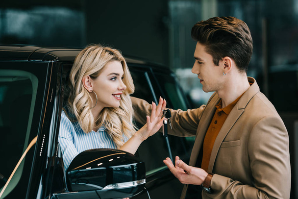 handsome man looking at cheerful blonde girl holding car key and sitting in automobile   - Фото, изображение