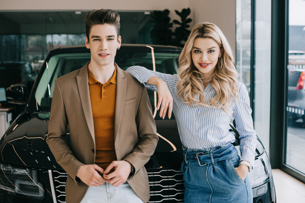handsome man and cheerful blonde girl standing with hand in pocket near car  - Fotografie, Obrázek