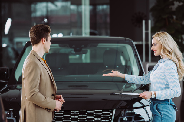 cheerful blonde car dealer showing automobile to handsome man in car showroom  - Photo, Image