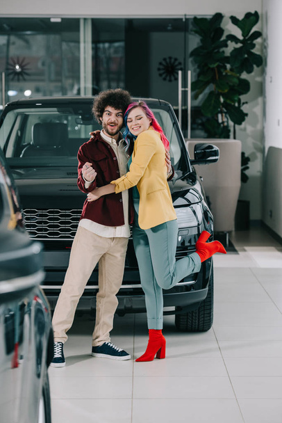 happy girl with colored hair hugging cheerful curly man in car showroom  - Photo, Image