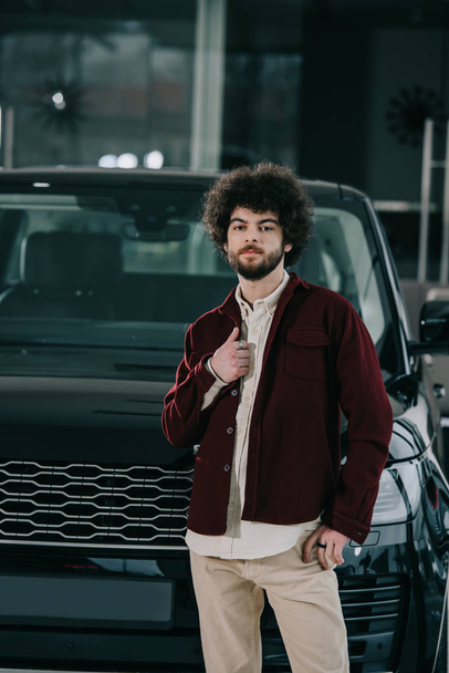 handsome bearded man standing near black automobile in car showroom  - Fotografie, Obrázek