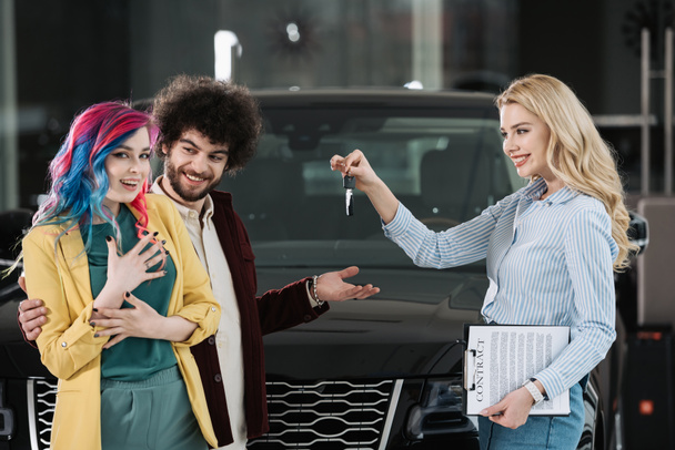 attractive blonde car dealer giving key to cheerful man and woman  - Фото, изображение