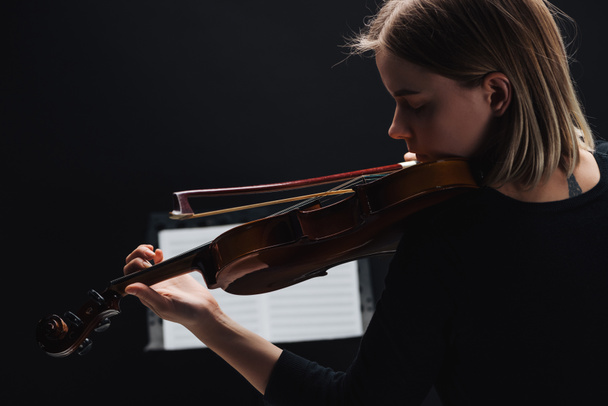 selective focus of young woman playing cello with bow in darkness with music book at background isolated on black  - Фото, изображение