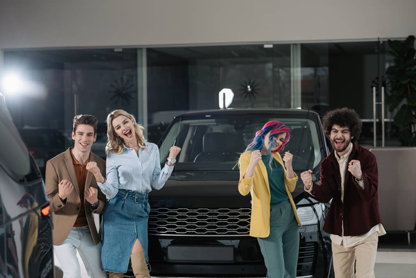 cheerful group of friends gesturing near automobile in car showroom  - Photo, Image