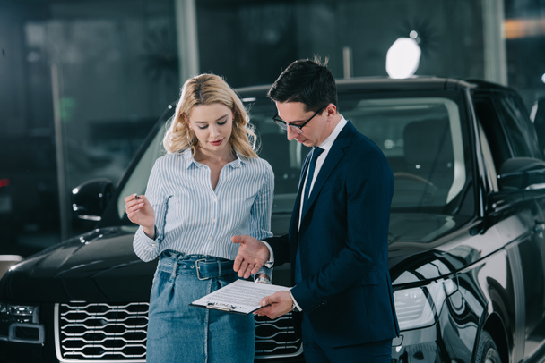 handsome car dealer holding clipboard near attractive blonde woman with pen  - Фото, изображение