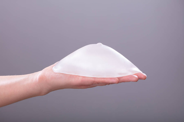 Close-up Of Woman's Hand Holding Silicon Breast Implant Against Gray Background - Foto, afbeelding