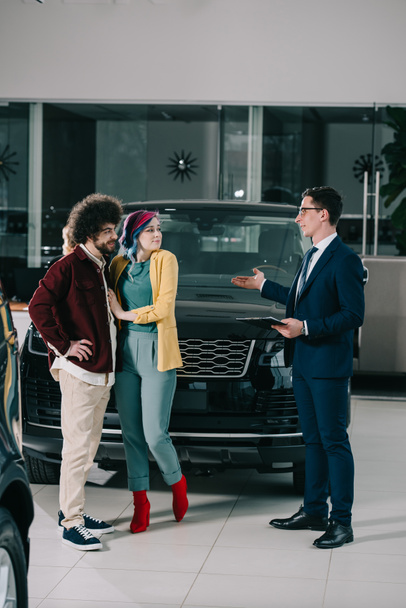 car dealer in glasses holding clipboard and gesturing near man and woman with colored hair  - Photo, Image