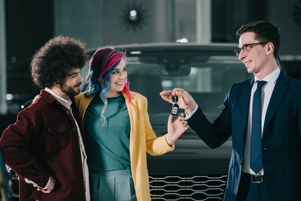 cheerful car dealer in glasses giving key to woman with colored hair and man  - Foto, Bild