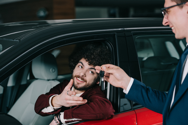 fröhlich lockiger Mann sitzt in rotem Auto und schaut auf Schlüssel in der Hand eines Autohändlers  - Foto, Bild