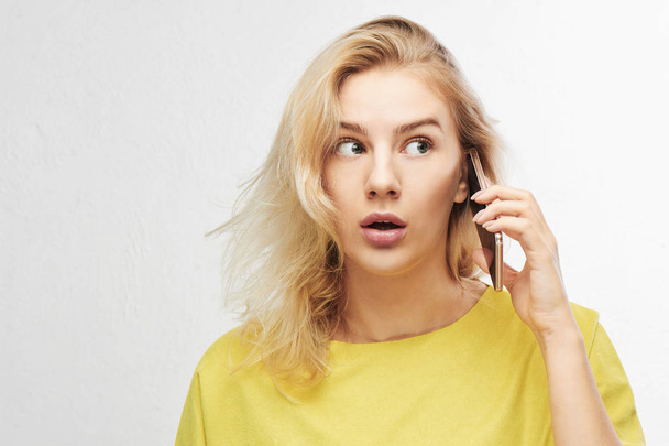 Portrait of shocked girl with open mouth talking mobile phone on white background with free advertising space. Woman in yellow casual outfit hears something amazing, she confused - Foto, Imagem