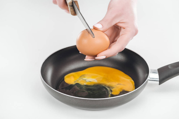 cropped view of woman smashing chicken egg with knife while preparing scrambled eggs in pan on white background - Foto, Bild