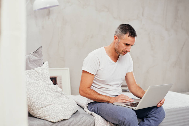 schöner kaukasischer Mann mittleren Alters lässig gekleidet mit Laptop, während er auf dem Bett im Schlafzimmer sitzt. - Foto, Bild