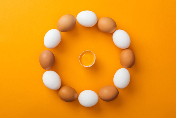 top view of white and brown organic chicken eggs arranged in round frame with smashed one inside on bright orange background  - Foto, imagen