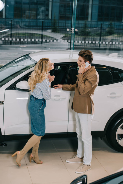 приваблива блондинка стоїть біля чоловіка, розмовляючи ом смартфон біля білої машини
 - Фото, зображення