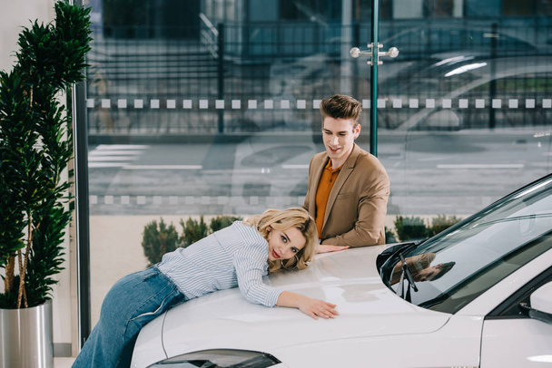 όμορφος άντρας κοιτάζοντας ελκυστική ξανθιά κορίτσι αγγίζοντας το λευκό αυτοκίνητο  - Φωτογραφία, εικόνα