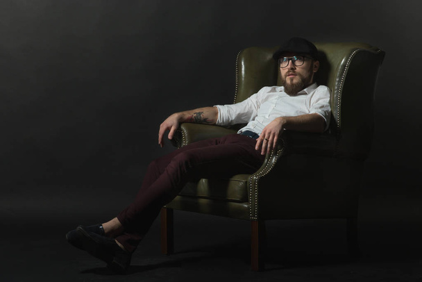 ein junger gutaussehender Mann mit Bart und Schnurrbart, schwarzer Mütze und weißem Hemd sitzt in einem ledernen grünen Sessel und zeigt Gesten mit den Händen in einem dunklen Studio - Foto, Bild