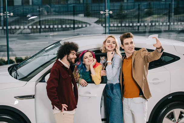 cheerful group of friends taking selfie on smartphone near white car  - 写真・画像