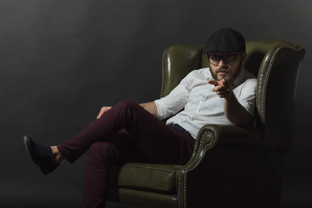 a young handsome man wearing beard and mustache in a black cap and a white shirt sitting in a leather green armchair conceived and showing gestures with his hands in a dark studio - Foto, imagen