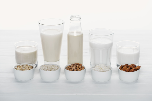 glasses and bottle with coconut, chickpea, oat, rice and almond milk on white wooden table with ingredients in bowls isolated on white - Fotó, kép