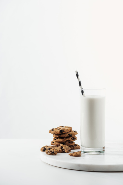 glass with fresh milk and straw near chocolate cookies on marble stand isolated on white - Фото, изображение
