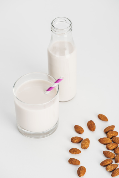 βιολογικό γάλα αμυγδάλου σε φιάλη και γυαλί με άχυρο κοντά σε διάσπαρτα αμύγδαλα - Φωτογραφία, εικόνα