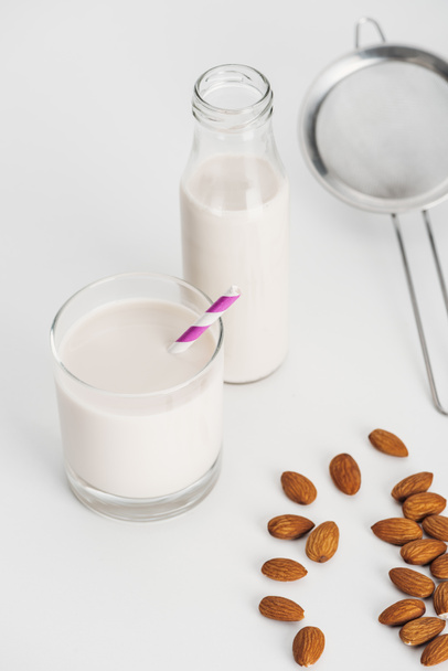βιολογικό γάλα αμυγδάλου σε φιάλη και γυαλί με άχυρο κοντά σε διάσπαρτα αμύγδαλα και κόσκινο - Φωτογραφία, εικόνα