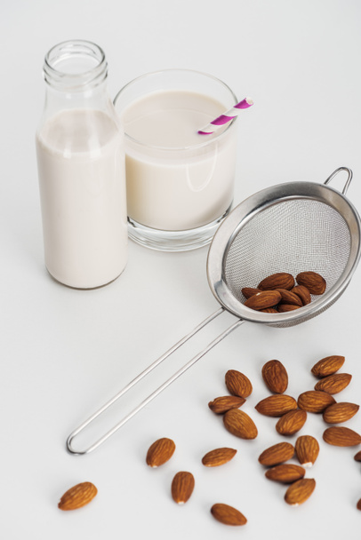 vegan almond milk in bottle and glass with straw near scattered almonds and sieve - Photo, Image