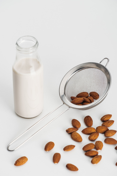 vegan almond milk in bottle near scattered almonds and sieve on grey background - Foto, afbeelding