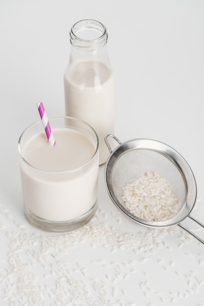 leite de arroz em garrafa e vidro com palha perto de arroz espalhado e peneira em fundo cinza
 - Foto, Imagem
