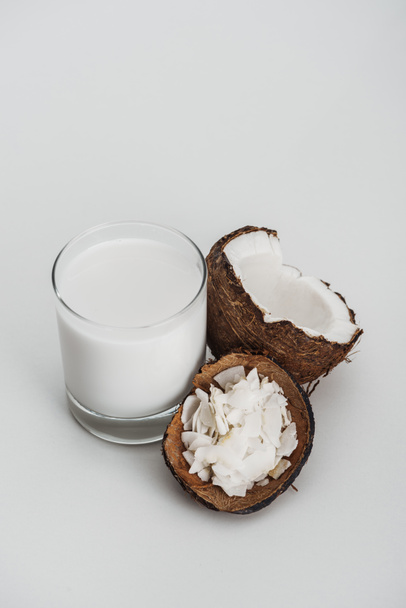 organische veganistische kokosmelk in glas in de buurt van kokos helft en chips op grijze achtergrond - Foto, afbeelding