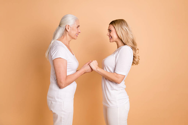 Profile side view portrait of nice-looking winsome attractive charming cheerful cheery ladies wearing white outfit holding hands trust support care generation isolated over beige pastel background - Photo, Image