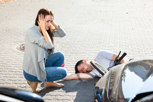 Junge Frau sieht bewusstlosen Radfahrer nach Unfall nahe Auto auf Straße liegen - Foto, Bild