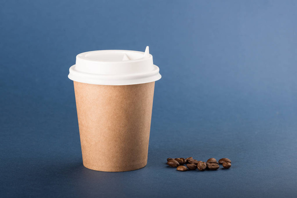 Kraft disposable cup with white cap and coffee beans on a blue background - Valokuva, kuva