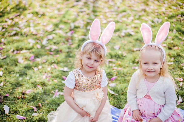 Niñas lindas están sentadas en la hierba cerca de la mogolia. Chicas con disfraces conejitos de Pascua
. - Foto, imagen