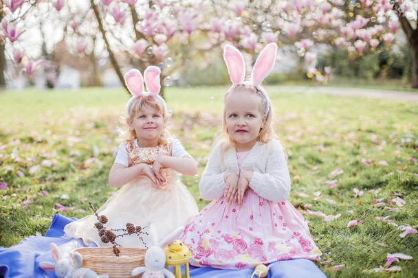 Kis aranyos lányok ülnek a füvön közelében mogolia. Lányok jelmezek húsvéti nyuszik. - Fotó, kép