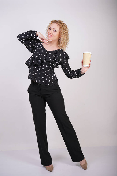 aranyos göndör szőke lány áll a fekete divatos ruhákat, és tart egy papírt csésze kávét egy fehér stúdió háttér - Fotó, kép