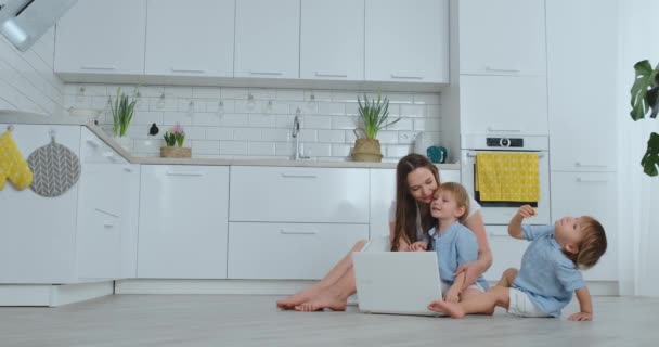 Tecnologia moderna. Moderno appartamento amorevole mamma e due piccoli figli seduti sul pavimento nel soggiorno guardare lo schermo del computer portatile. I bambini con mamma giocano su un computer portatile
. - Filmati, video