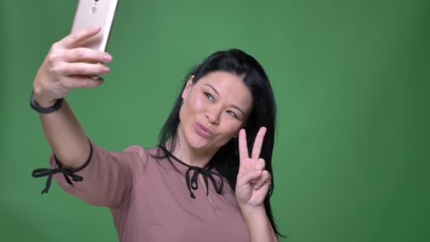 Closeup shoot of young attractive asian female with black hair taking selfies on the the phone and posing in front of the camera with background isolated on green - Záběry, video