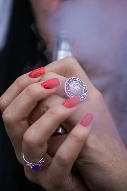 Mujer caucásica con uñas rojas manicura y anillo antiguo en el dedo sostiene pequeño vapor. Fumar vay alternativa. La mujer exhala humo espeso. La vida sin cigarrillos. Mujer-vaper. Pequeño cigarrillo electrónico. - Foto, Imagen
